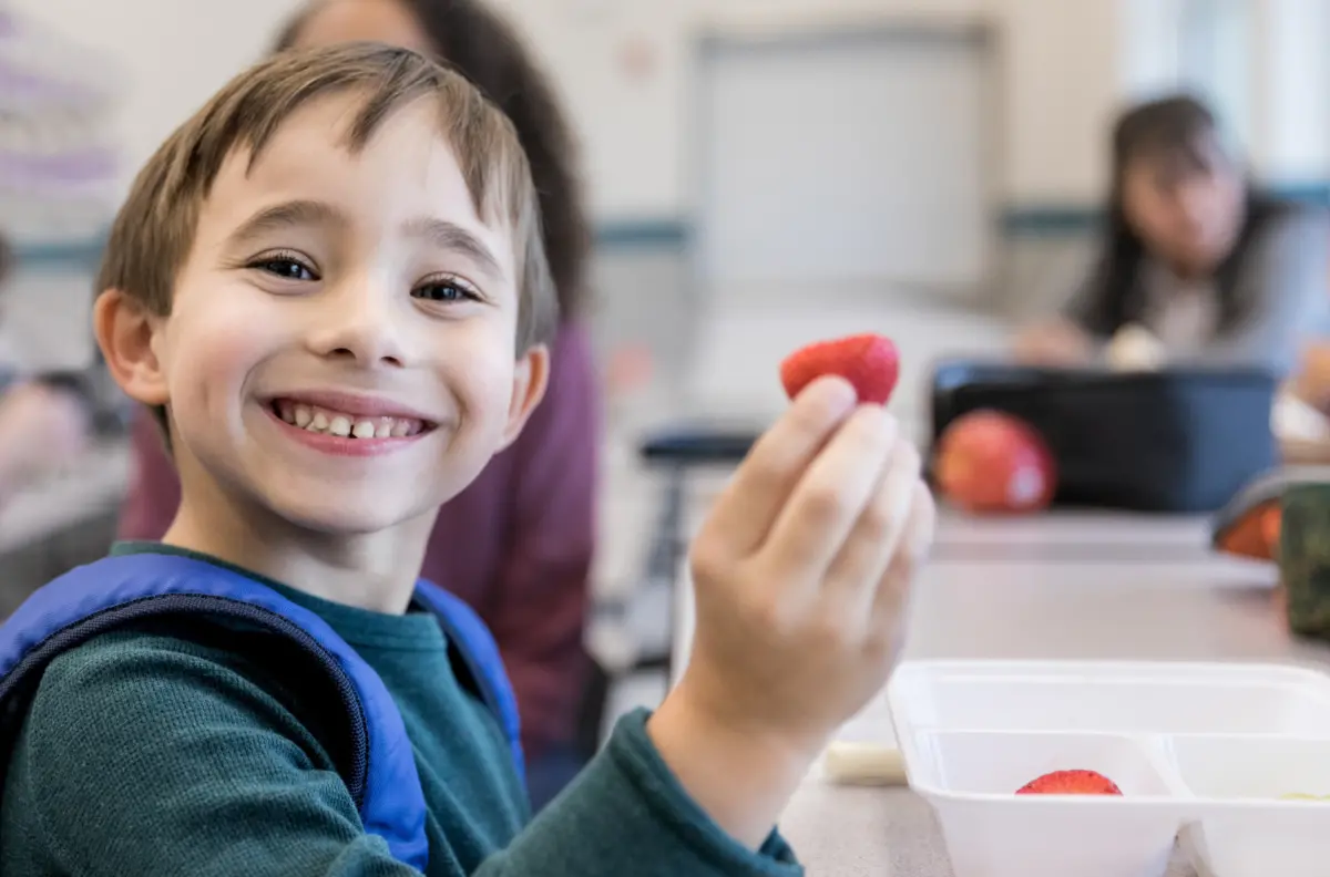 A young boy smiles and holds up a strawberry in a school cafeteria