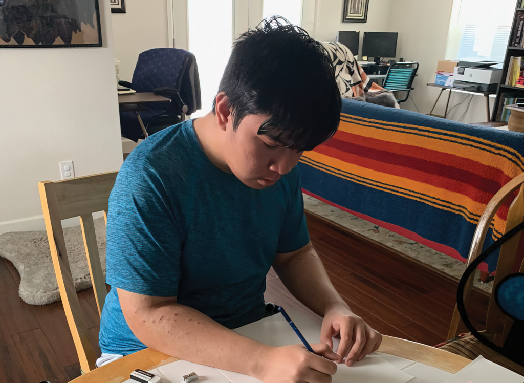 A teenage boy sits writing at a table in his home